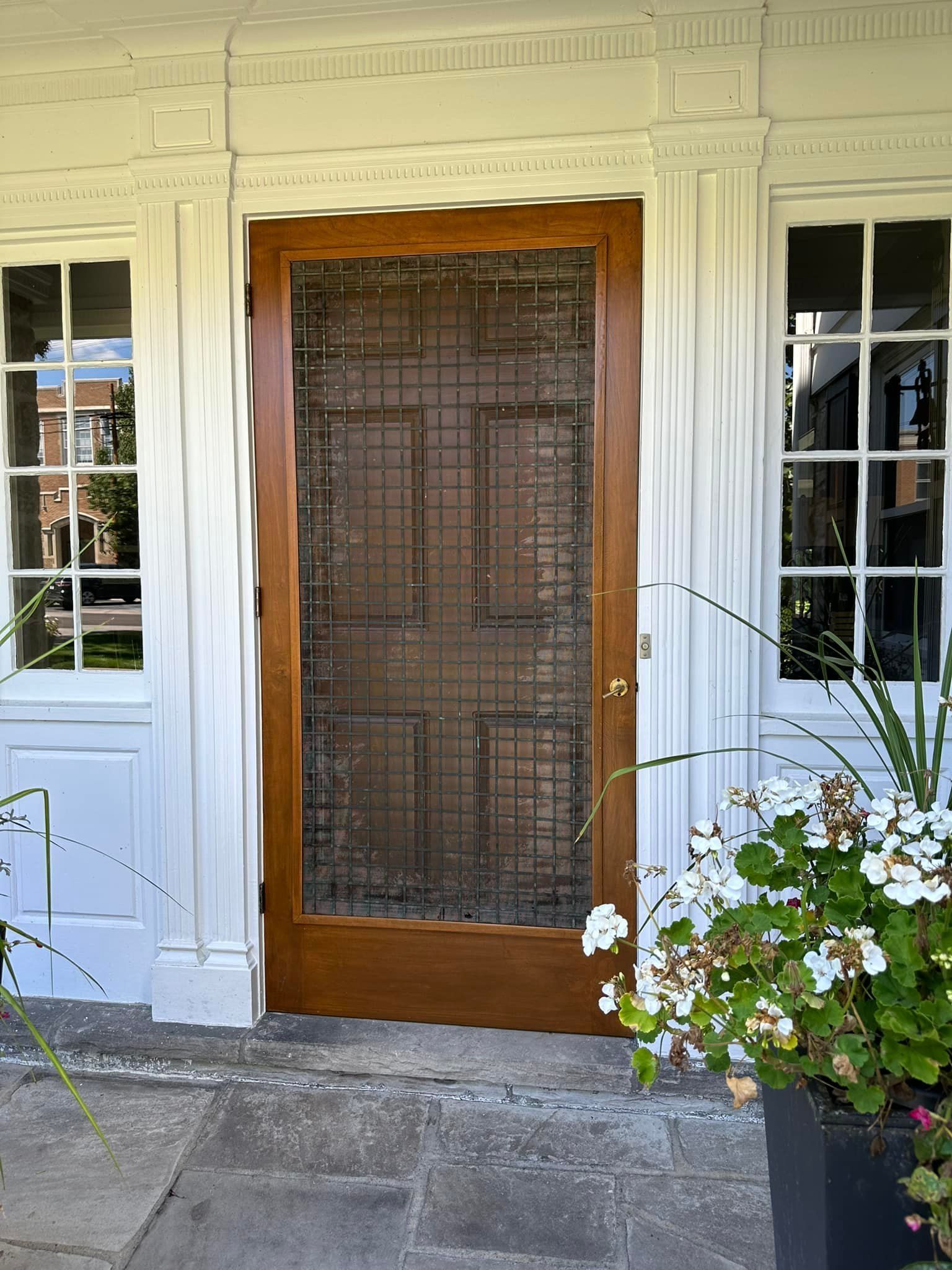 Wooden door with screen, flanked by white columns and windows, with potted flowers nearby.