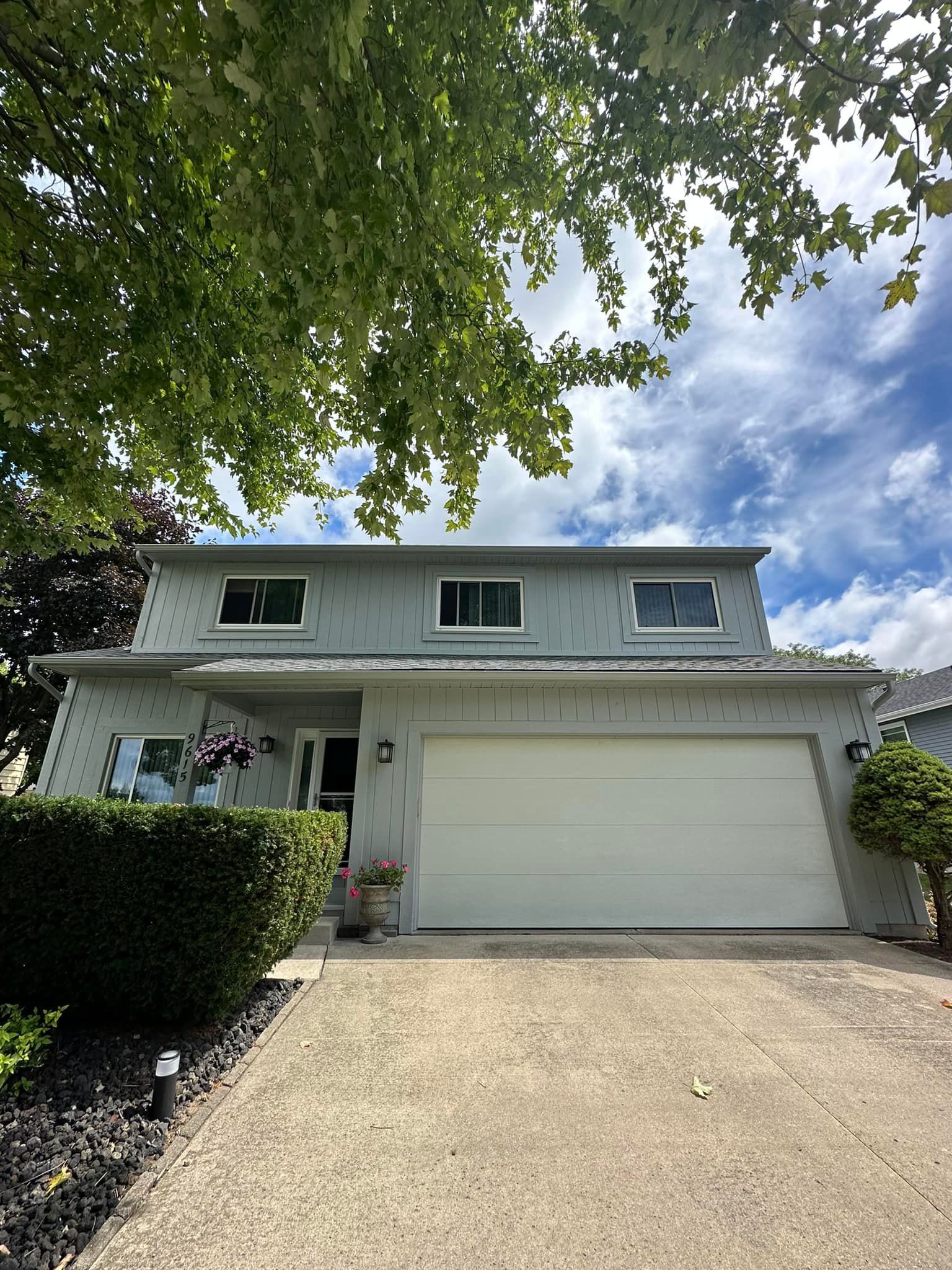 Two-story house with gray siding, white garage door, and green foliage against a blue sky.
