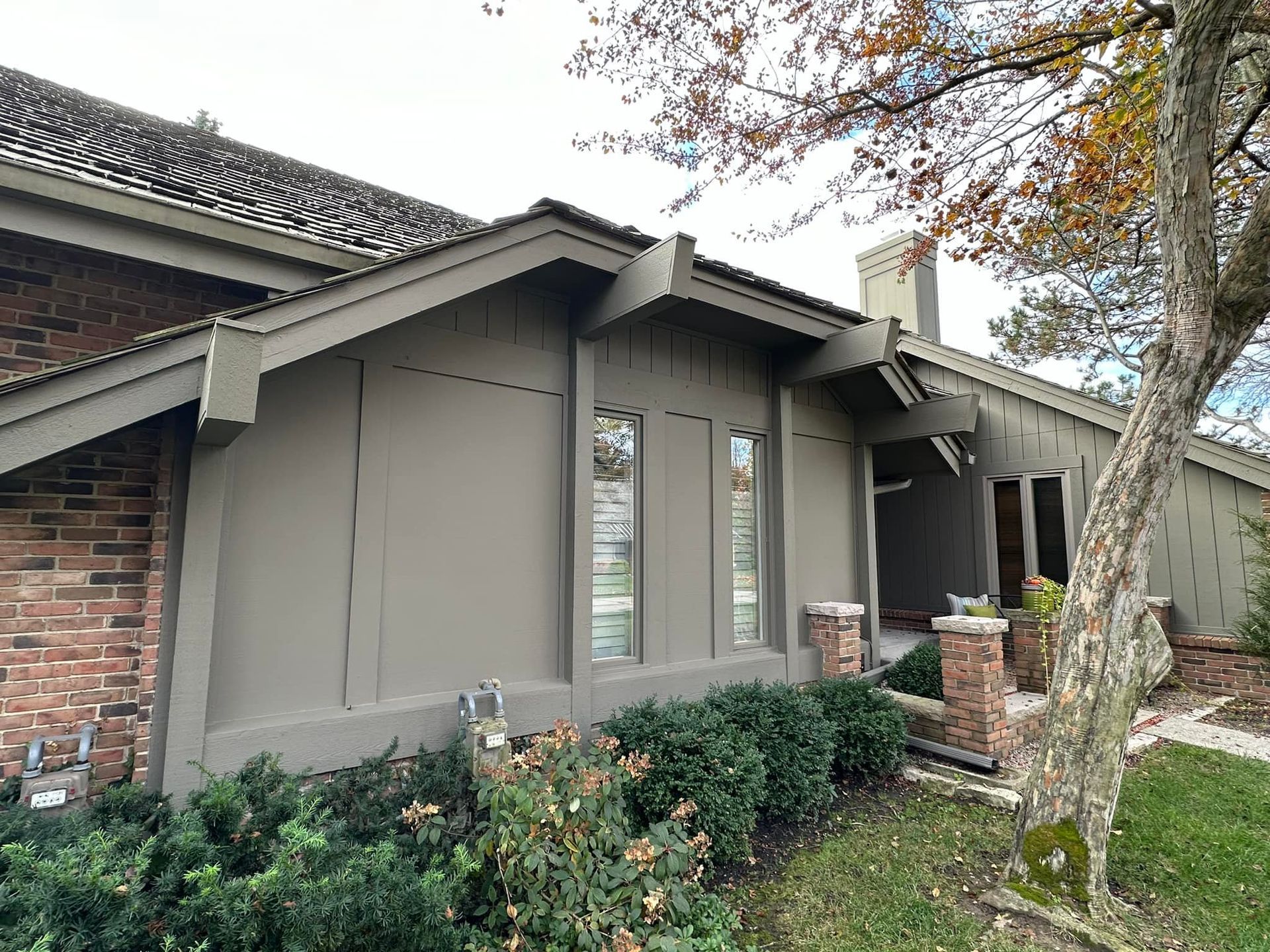 A beige-painted house with three vertical windows, brick accents, and a tree in the foreground.