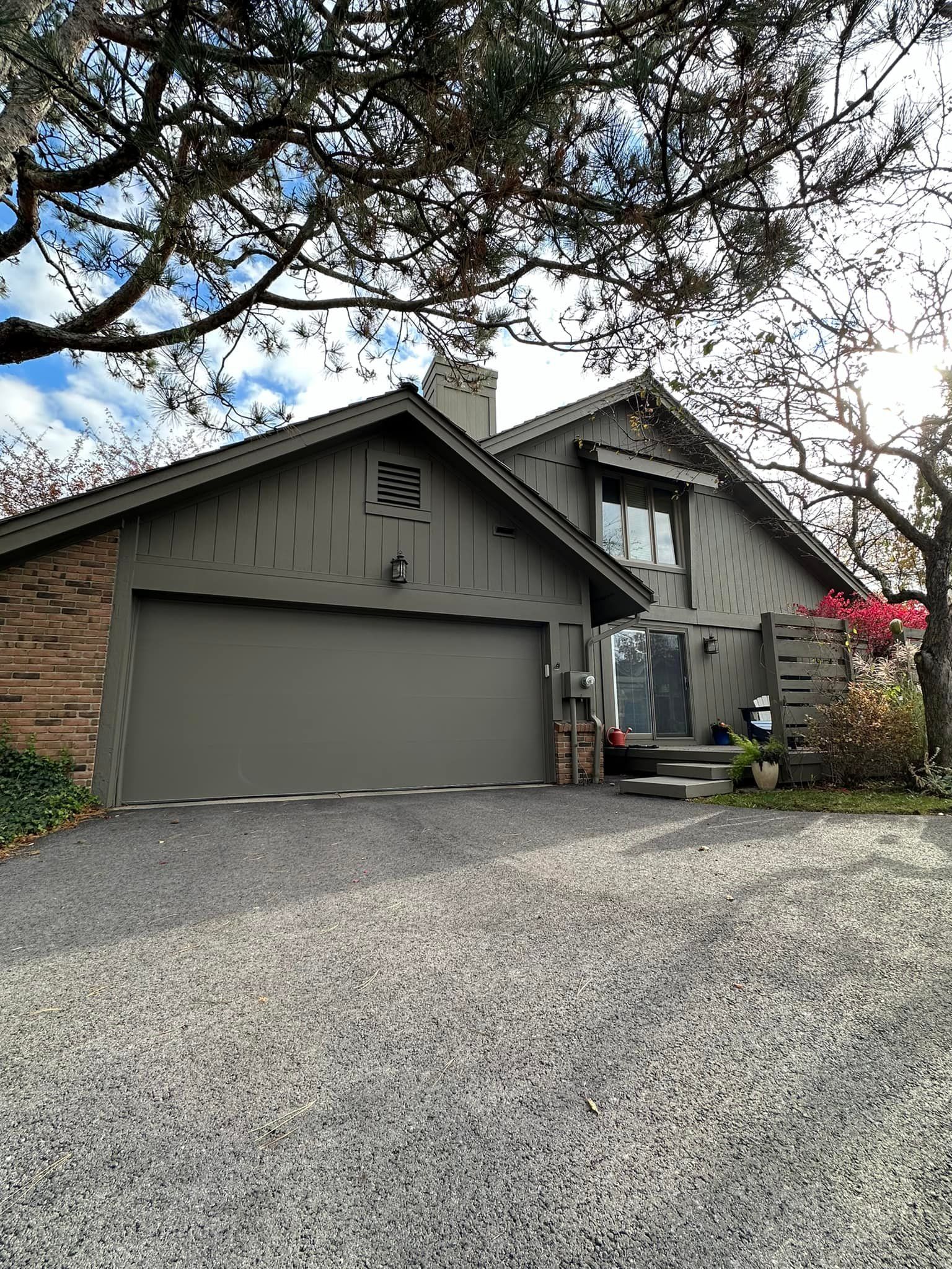 House with gray siding and garage, brick accents, gravel driveway, under tree branches.