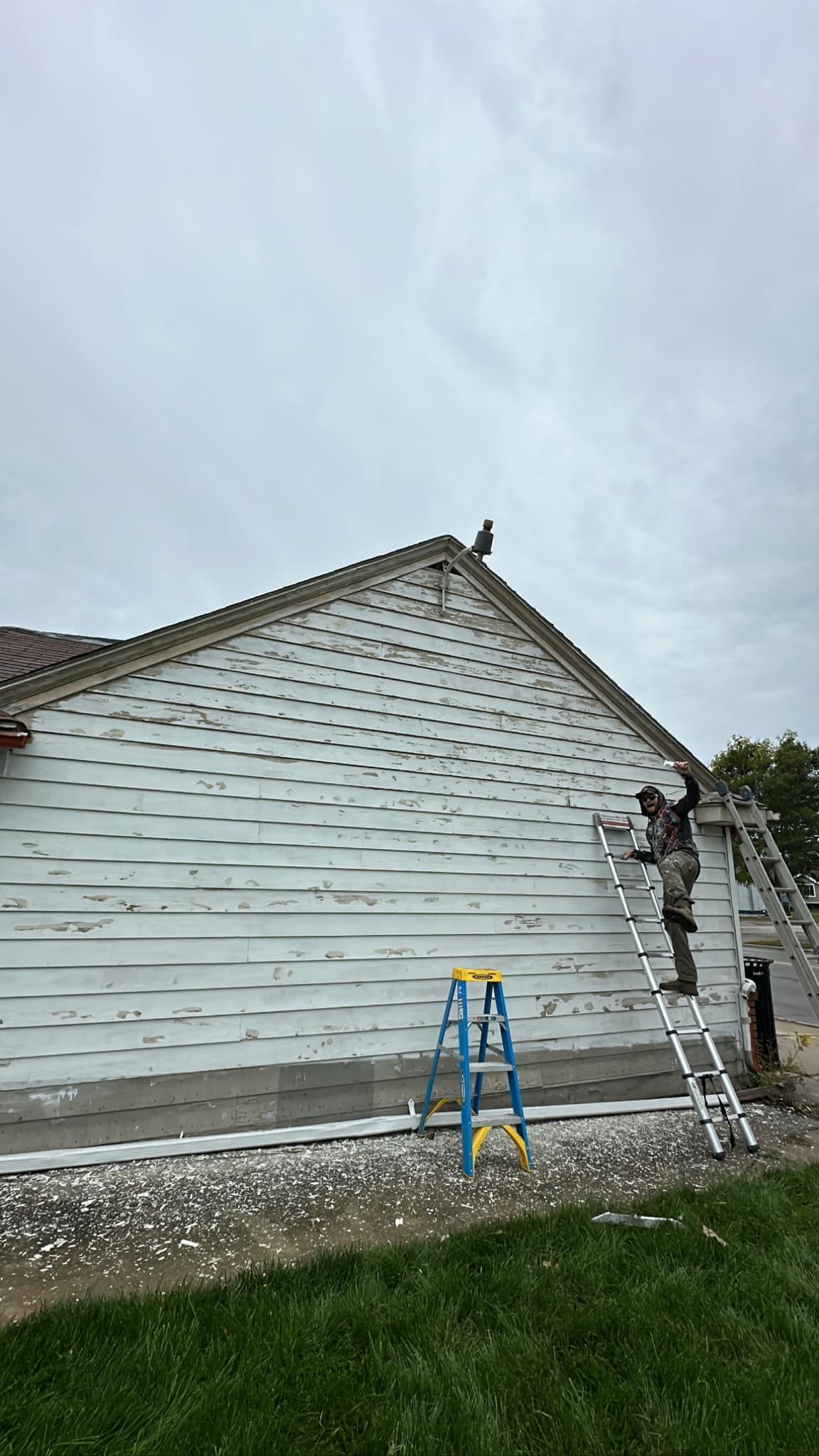 Workers on ladders painting the side of a white house with a gray roof on an overcast day.