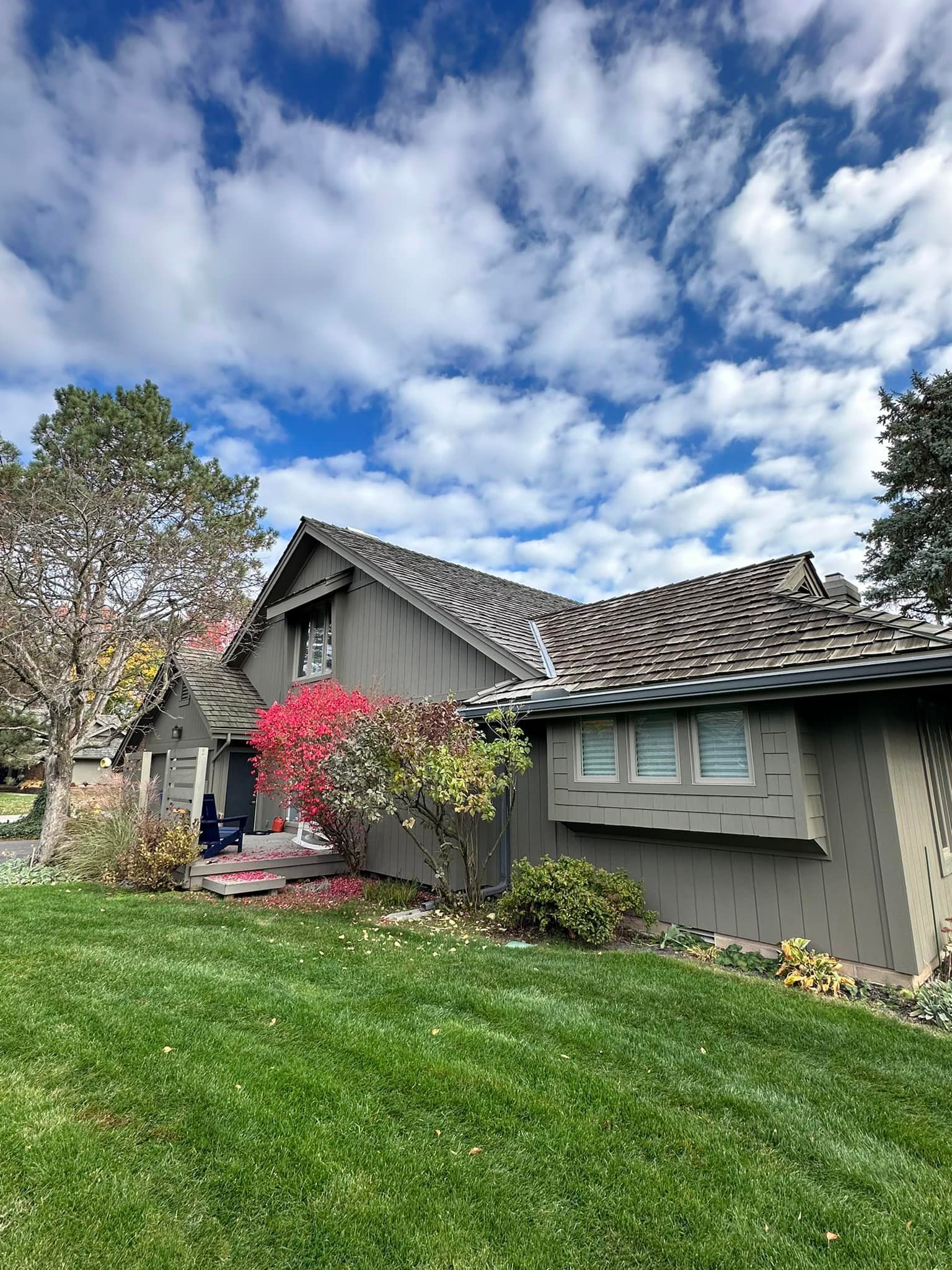 House with brown siding, green lawn, autumn foliage, and blue sky with clouds.