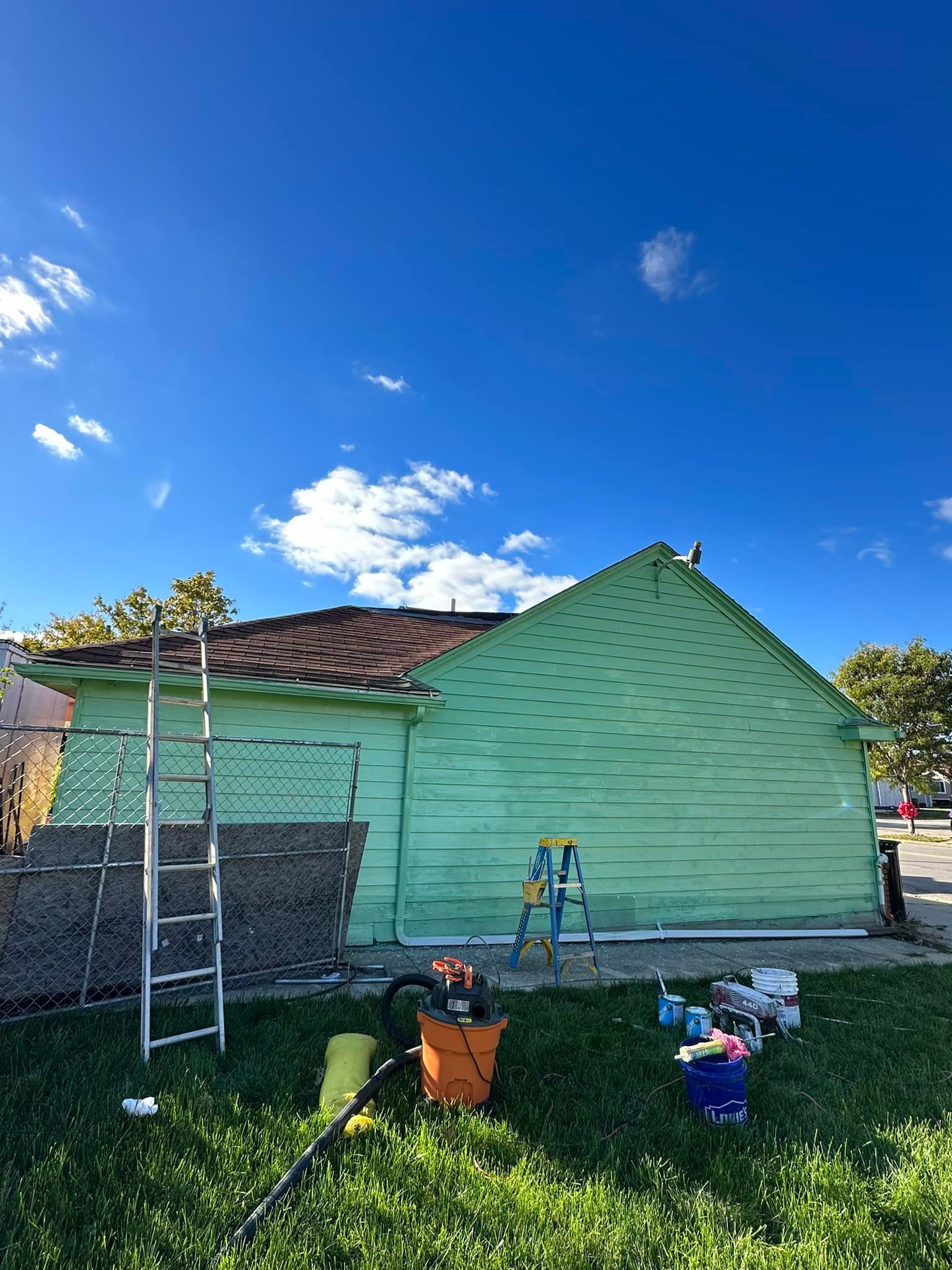 Green house being painted, ladder, tools on lawn, blue sky.