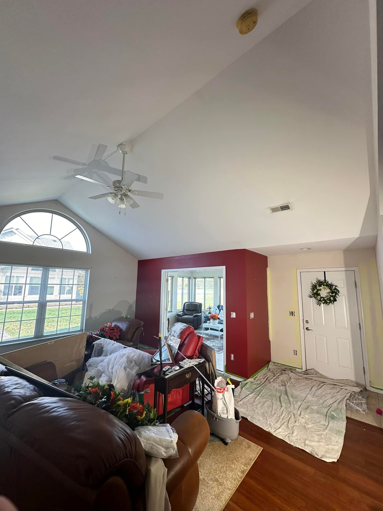 Living room with vaulted ceiling, large arched window, red accent wall, white door with wreath, and cluttered furniture.
