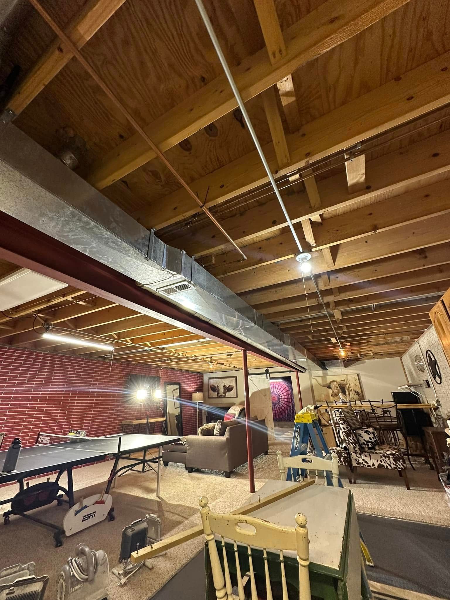 Basement entertainment area with ping pong table, couch, and chair under exposed wood beams and ductwork.