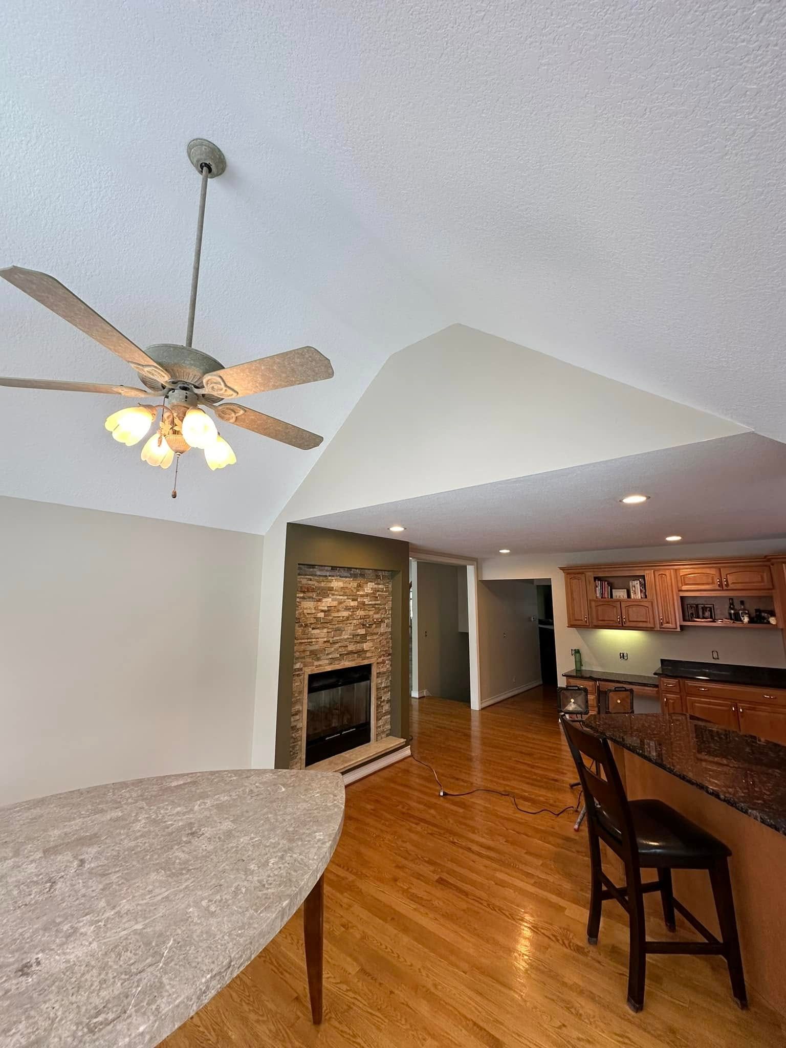 Interior shot of living space with fireplace, kitchen, and ceiling fan. Light wood flooring.
