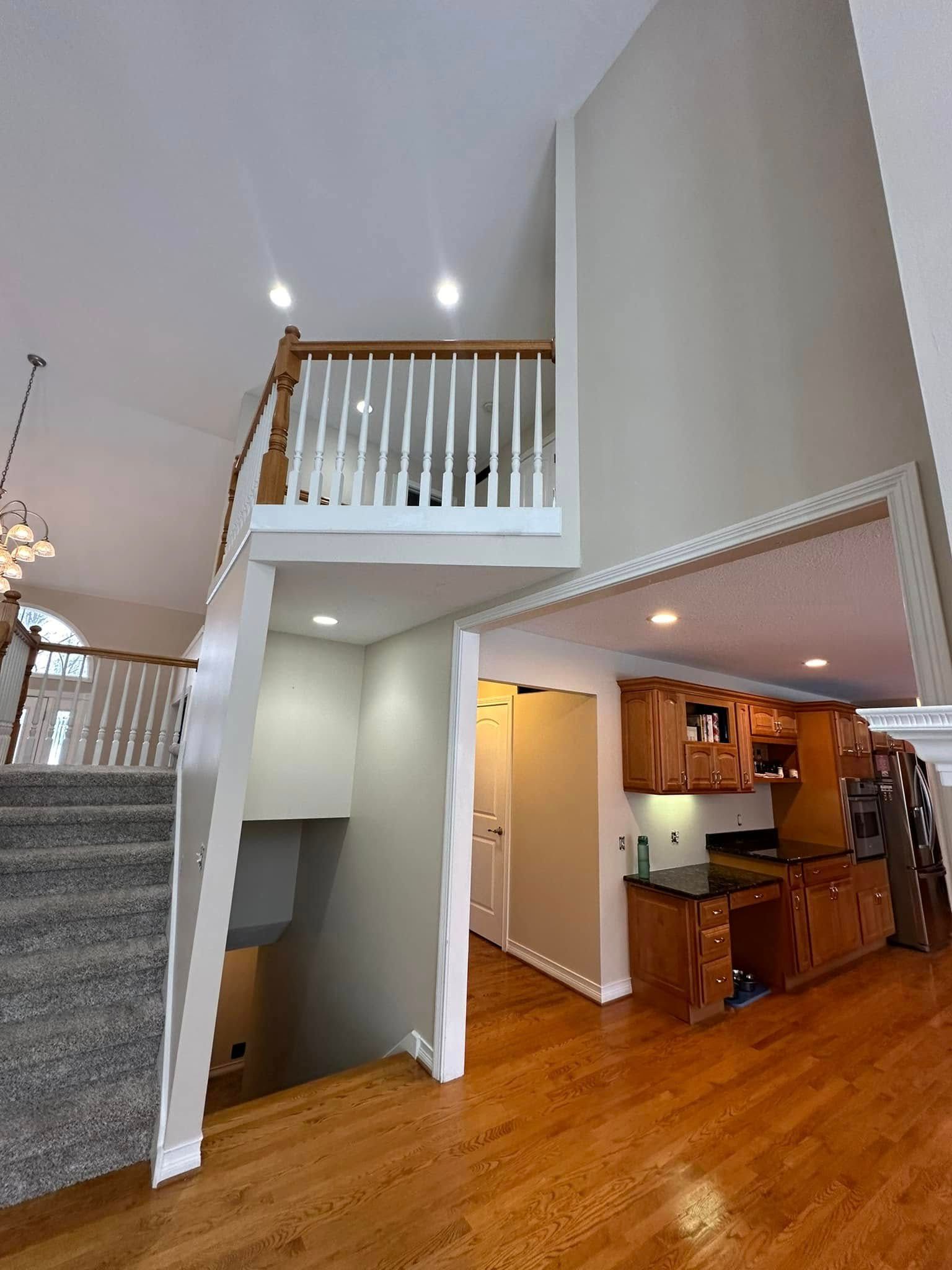 Interior view of a house with a staircase, kitchen, and loft. Wooden floors and cabinets. Gray carpeted stairs.