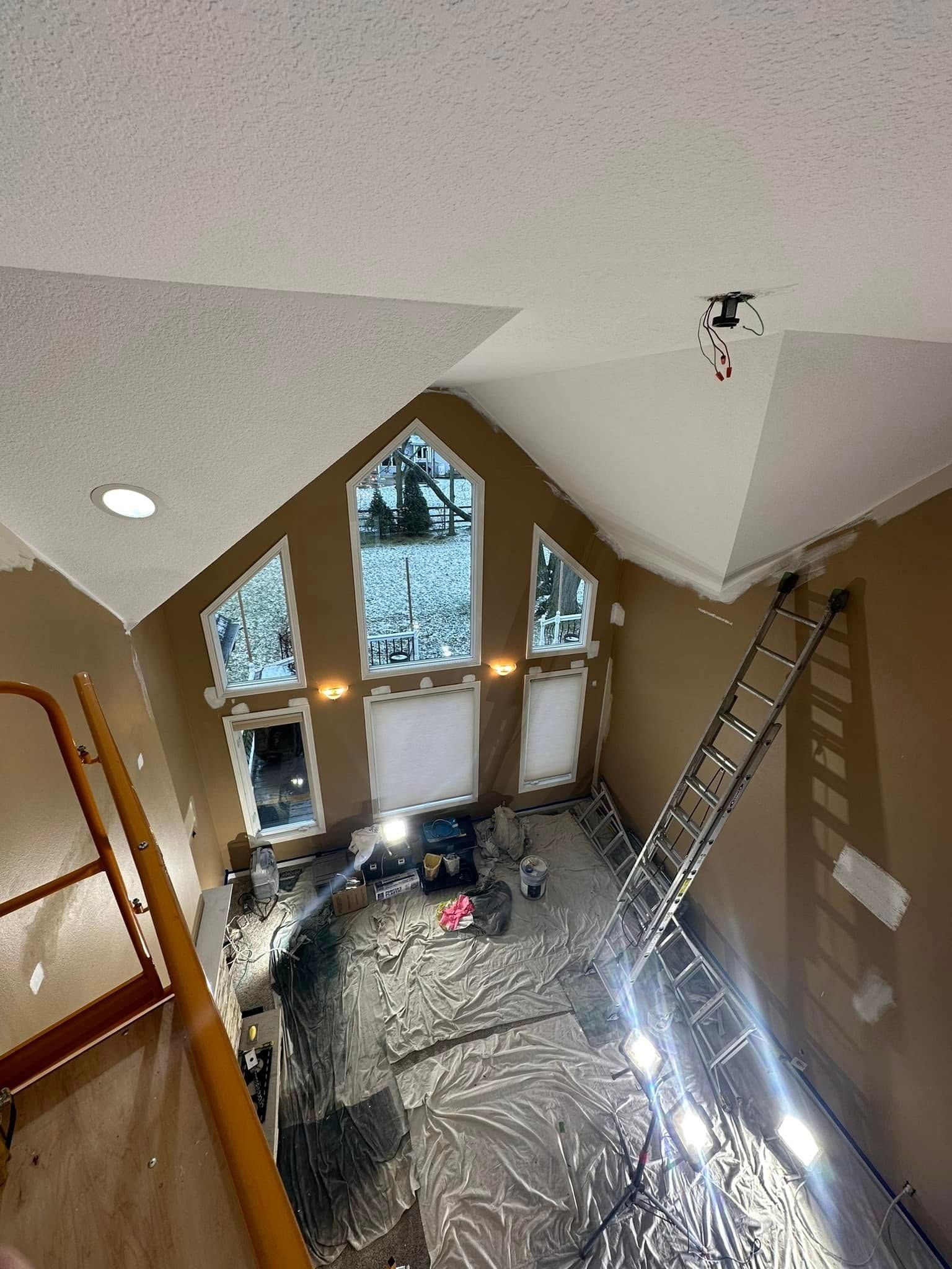 Interior view of a room with high ceilings. A ladder stands by a large window with plastic sheeting on the floor.