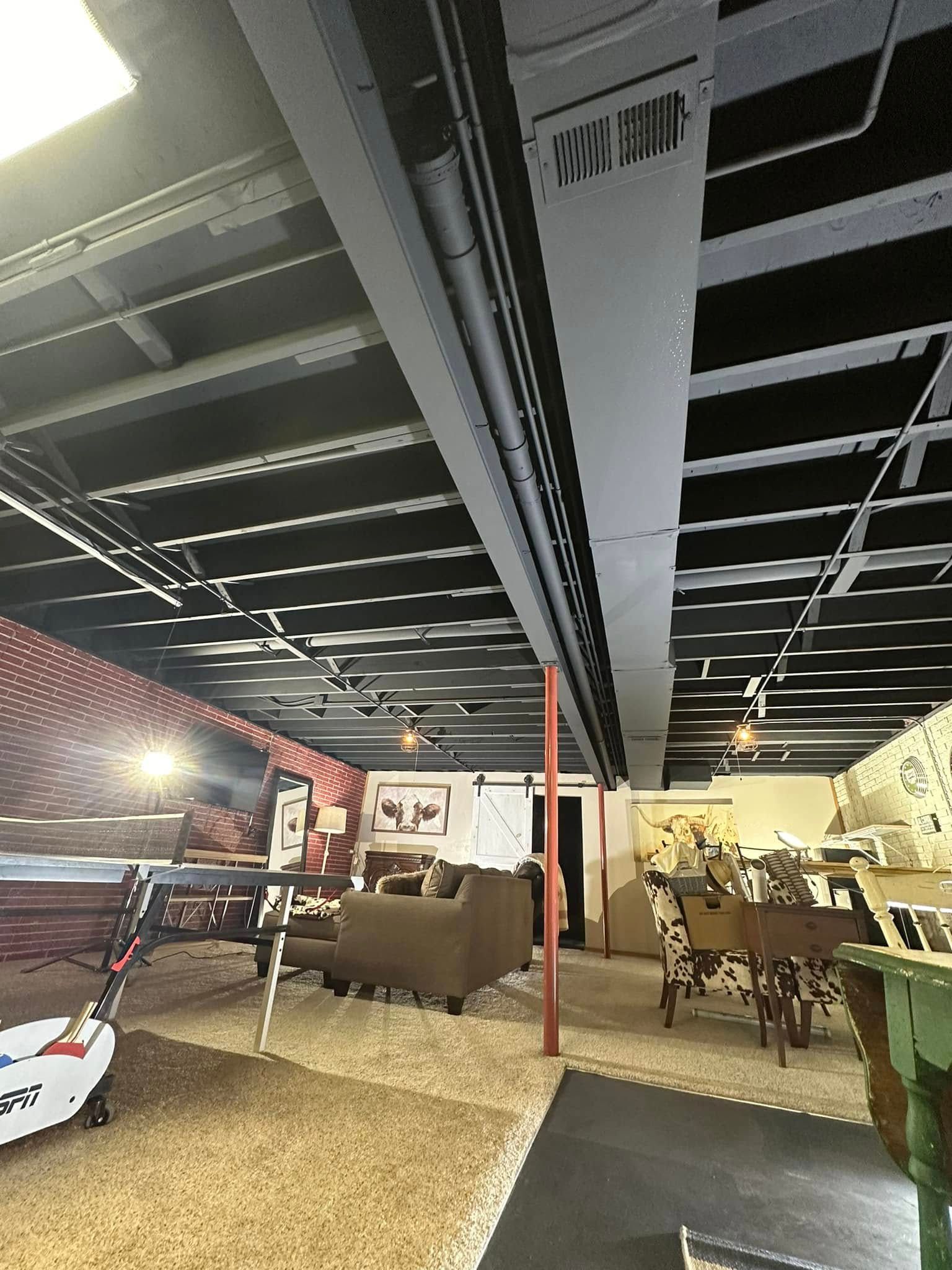 Basement ceiling with HVAC ductwork, black beams, and a glimpse of a recreation area with furniture.