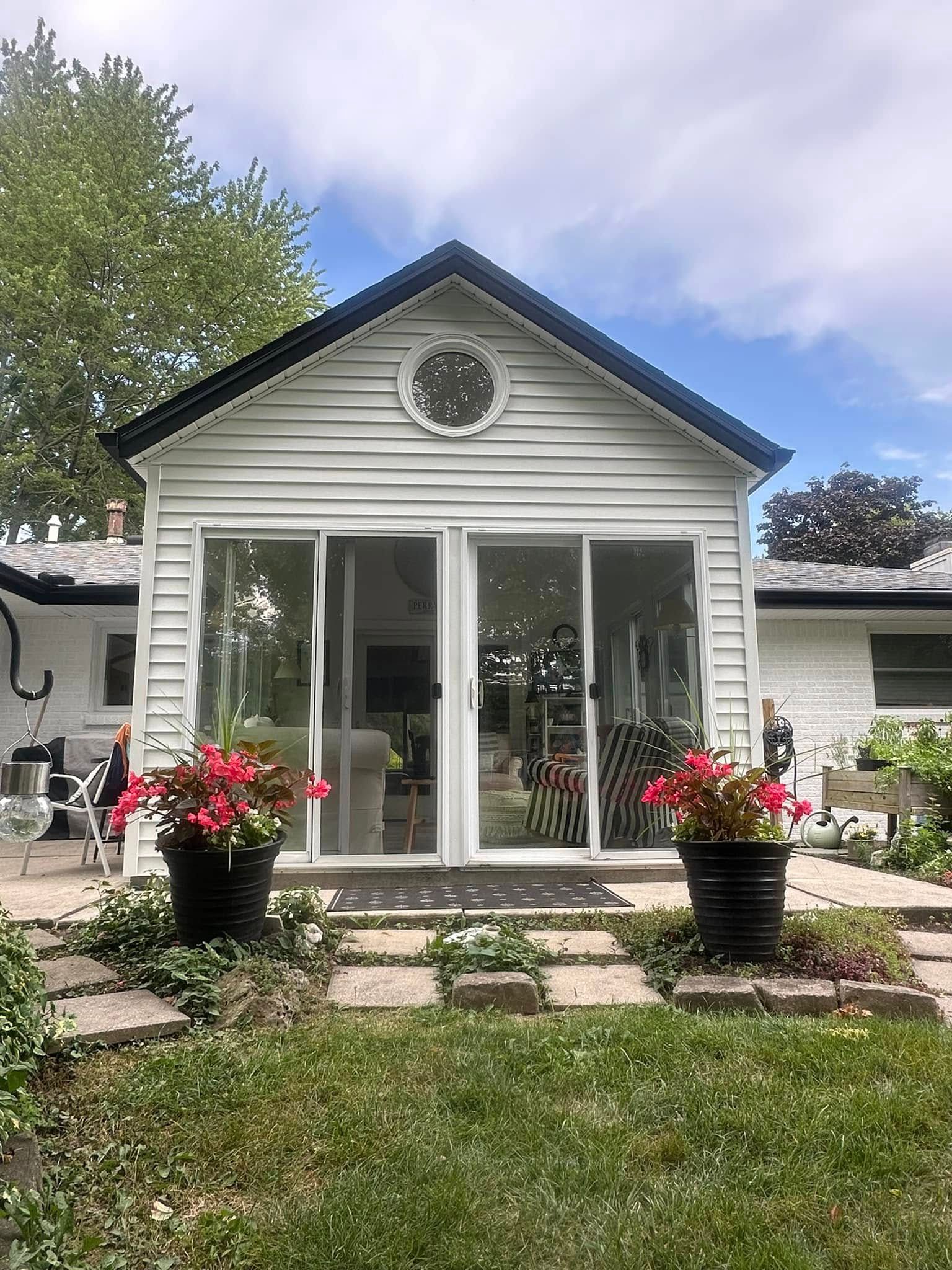 White-sided sunroom with glass doors, black roof, flower pots, and stone pathway leading from the grass.