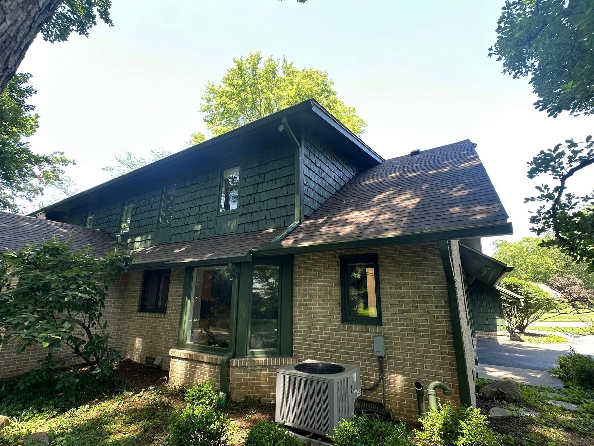 Green-sided house with brick base, brown roof, and air conditioner unit. Surrounded by trees and greenery.
