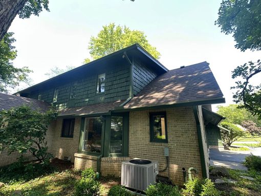 Green-sided house with brick base, brown roof, and air conditioner unit. Surrounded by trees and greenery.