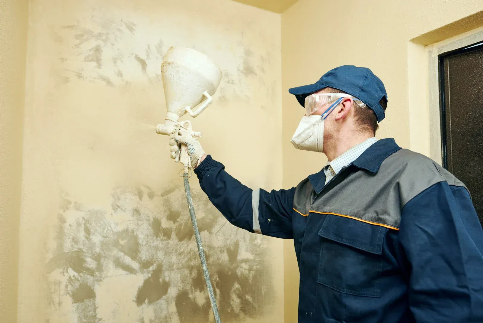 Man in protective gear sprays textured material on a wall indoors.