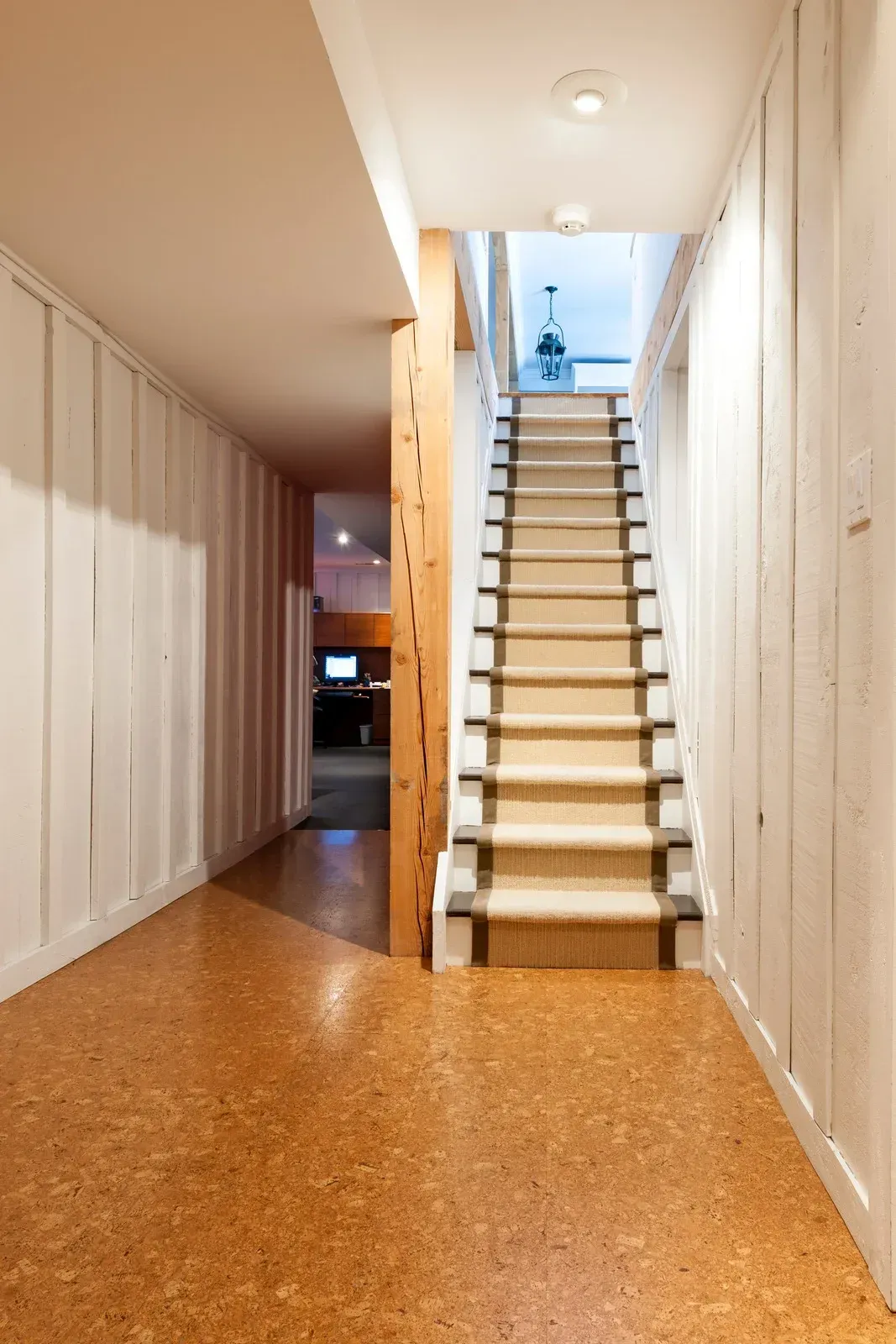 Hallway with stairs and cork flooring, white paneled walls, and a wood support beam.