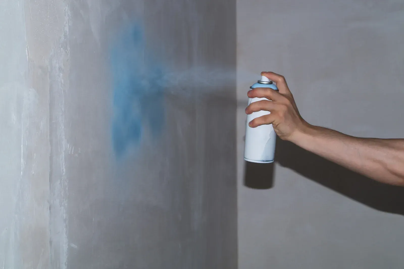 Hand spraying blue paint onto a gray wall in a corner.