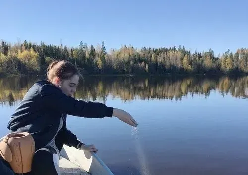 Woman in boat scattering contents over water, trees and blue sky in background.