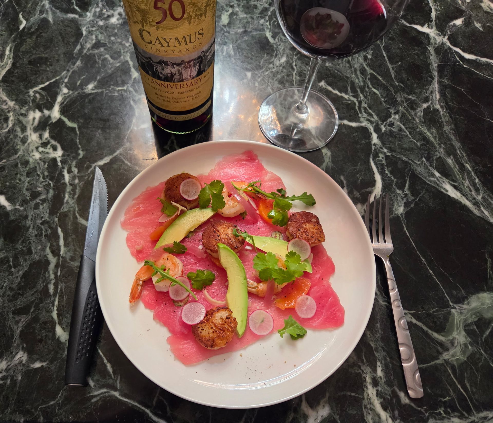 Plate with tuna carpaccio, fried scallops, avocado, wine bottle and glass, and cutlery on a marble surface.
