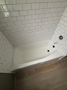 White bathtub surrounded by white subway tile and wood-look flooring. Chrome shower faucet.
