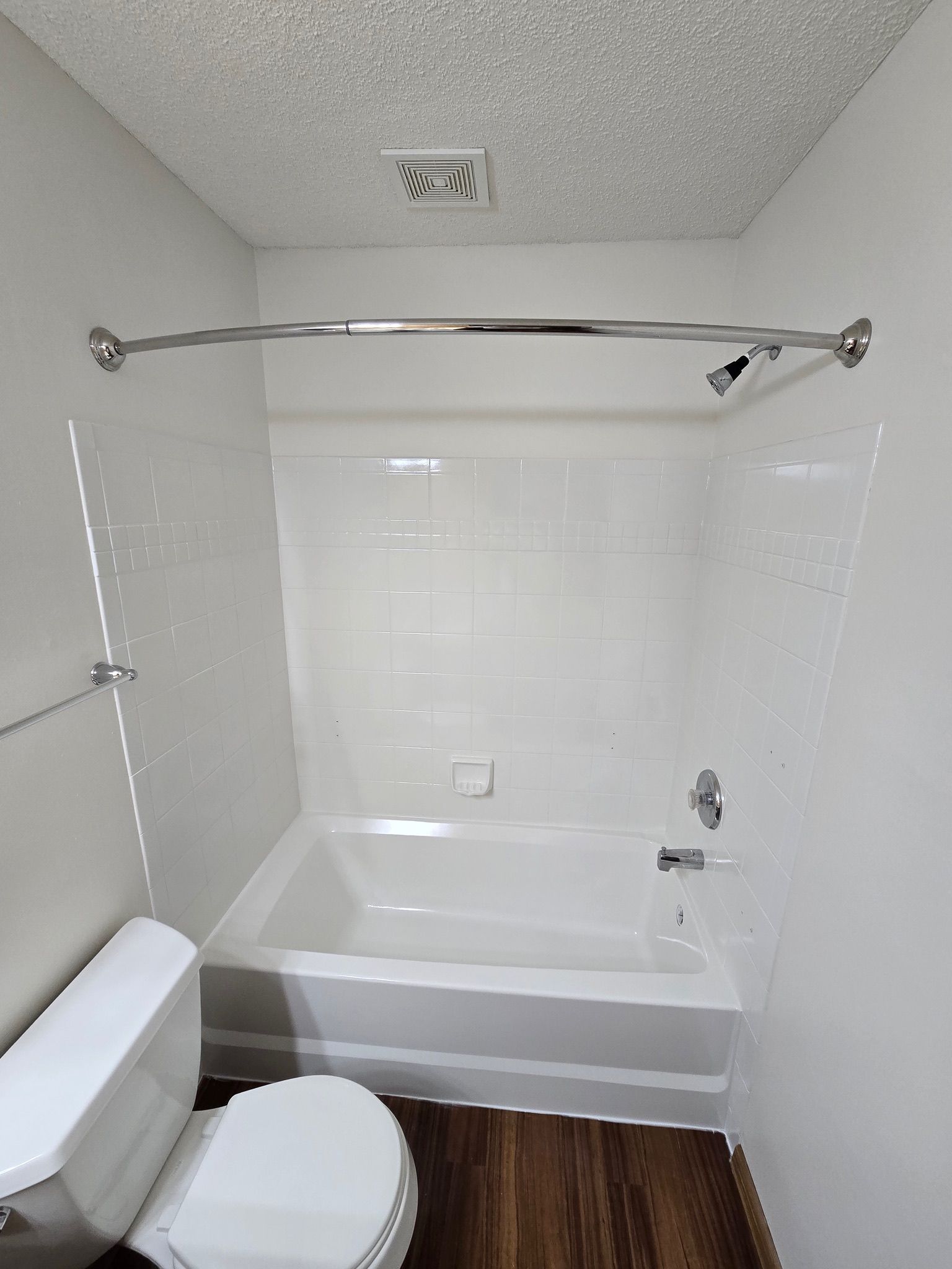 Bathroom with white tub, shower rod, and toilet; brown floor, white walls and ceiling.