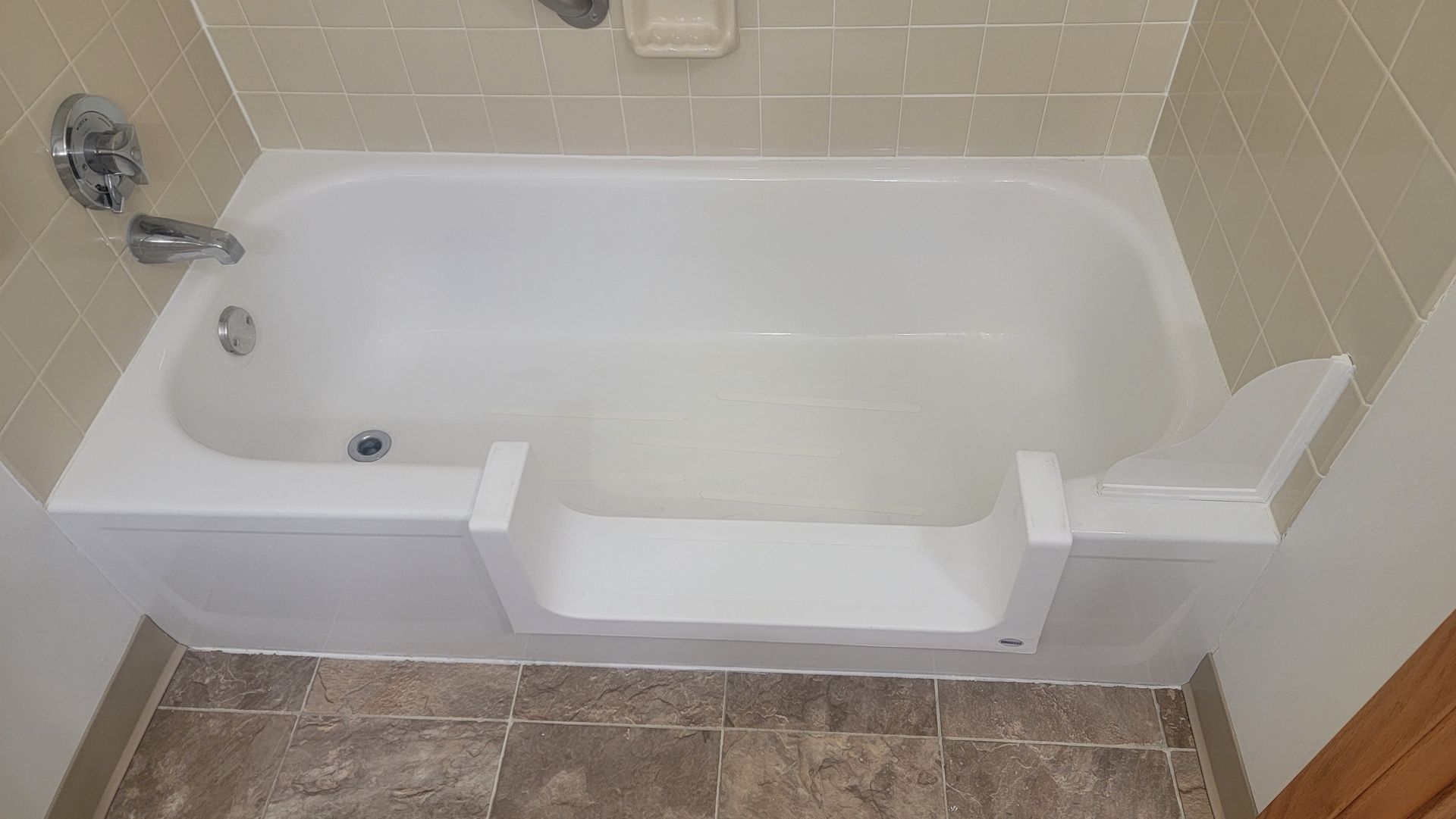 White walk-in bathtub with step, chrome fixtures, beige tile surround, and brown tile floor.
