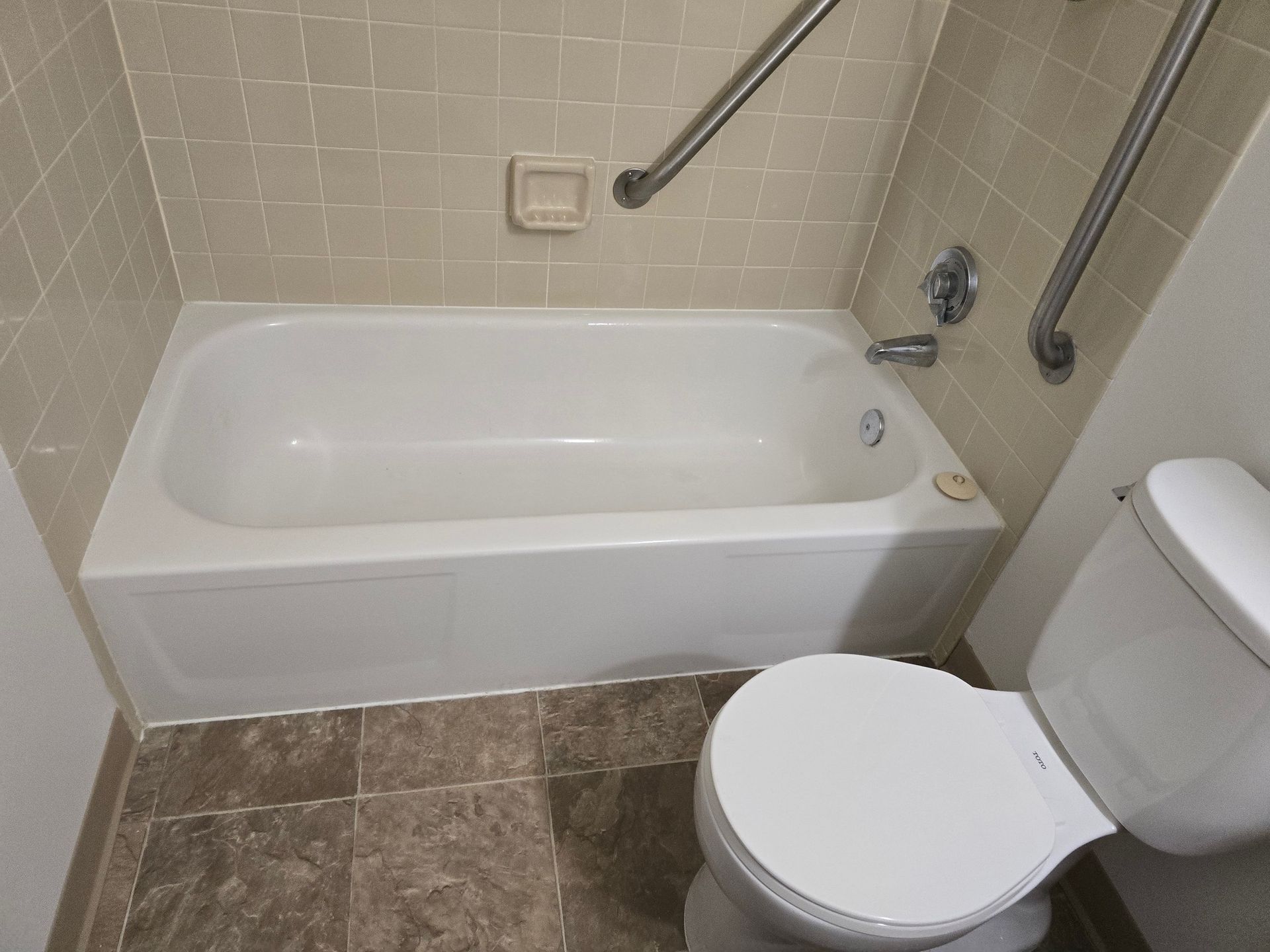 Bathtub and toilet in bathroom with tile walls, grab bars, and brown floor tiles.