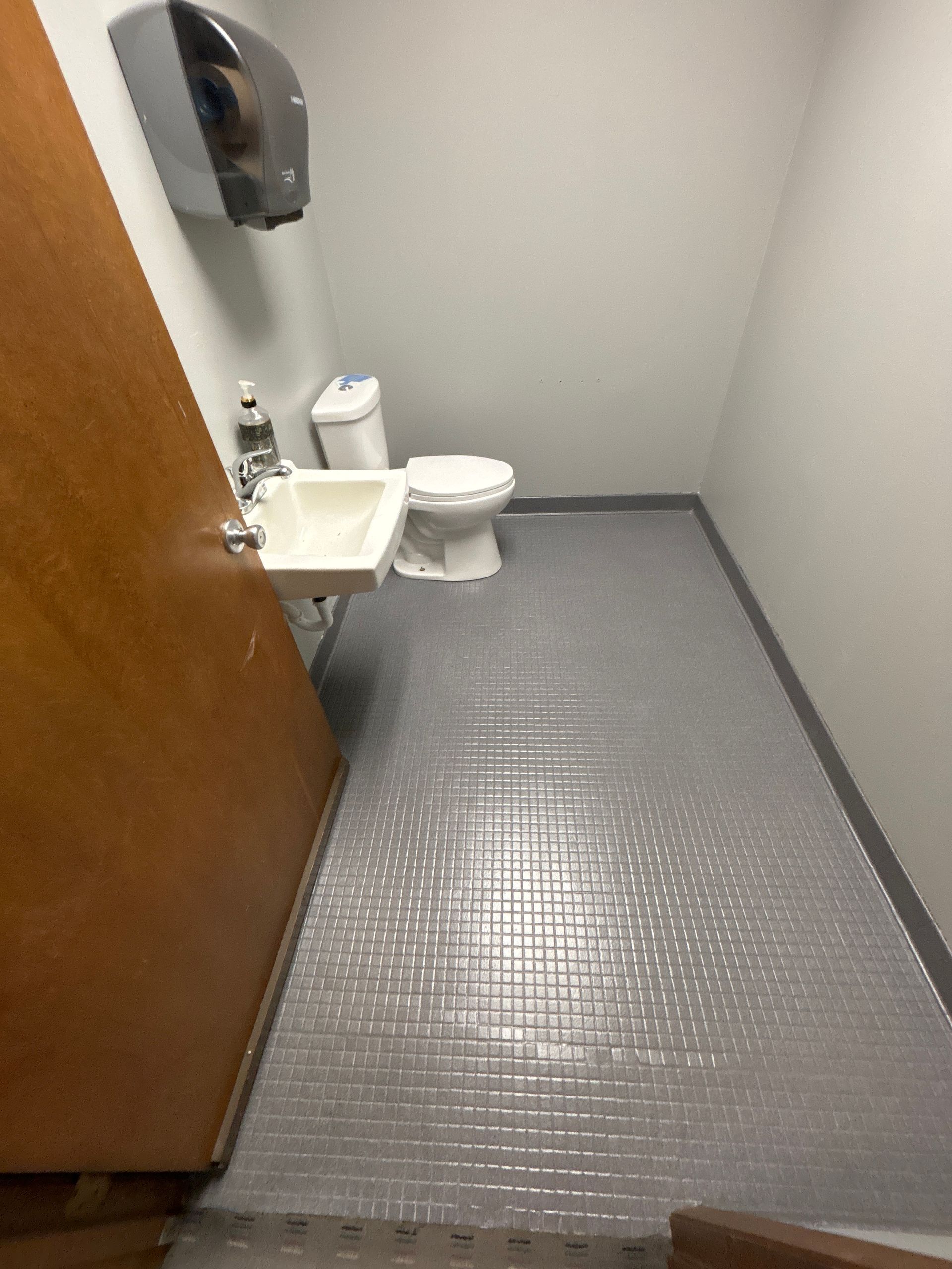 Small public restroom with gray tiled floor, toilet, sink, and paper towel dispenser.