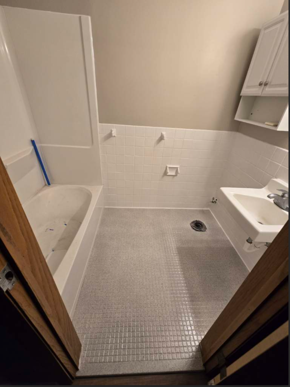 Small bathroom with white tiled walls, bathtub, and sink. Gray tile flooring. Brown door frame.