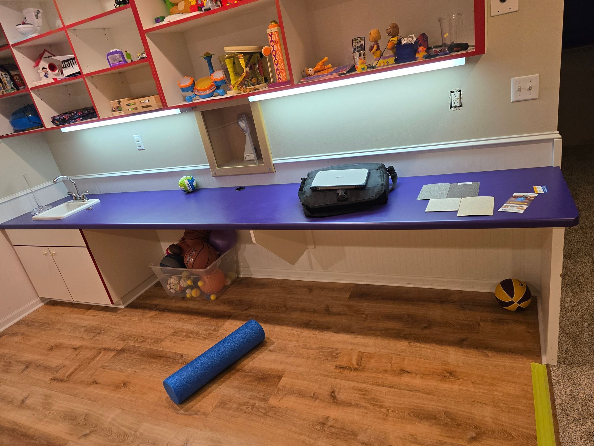 Playroom with built-in desk, storage shelves, sports equipment, and a foam roller on wooden flooring.