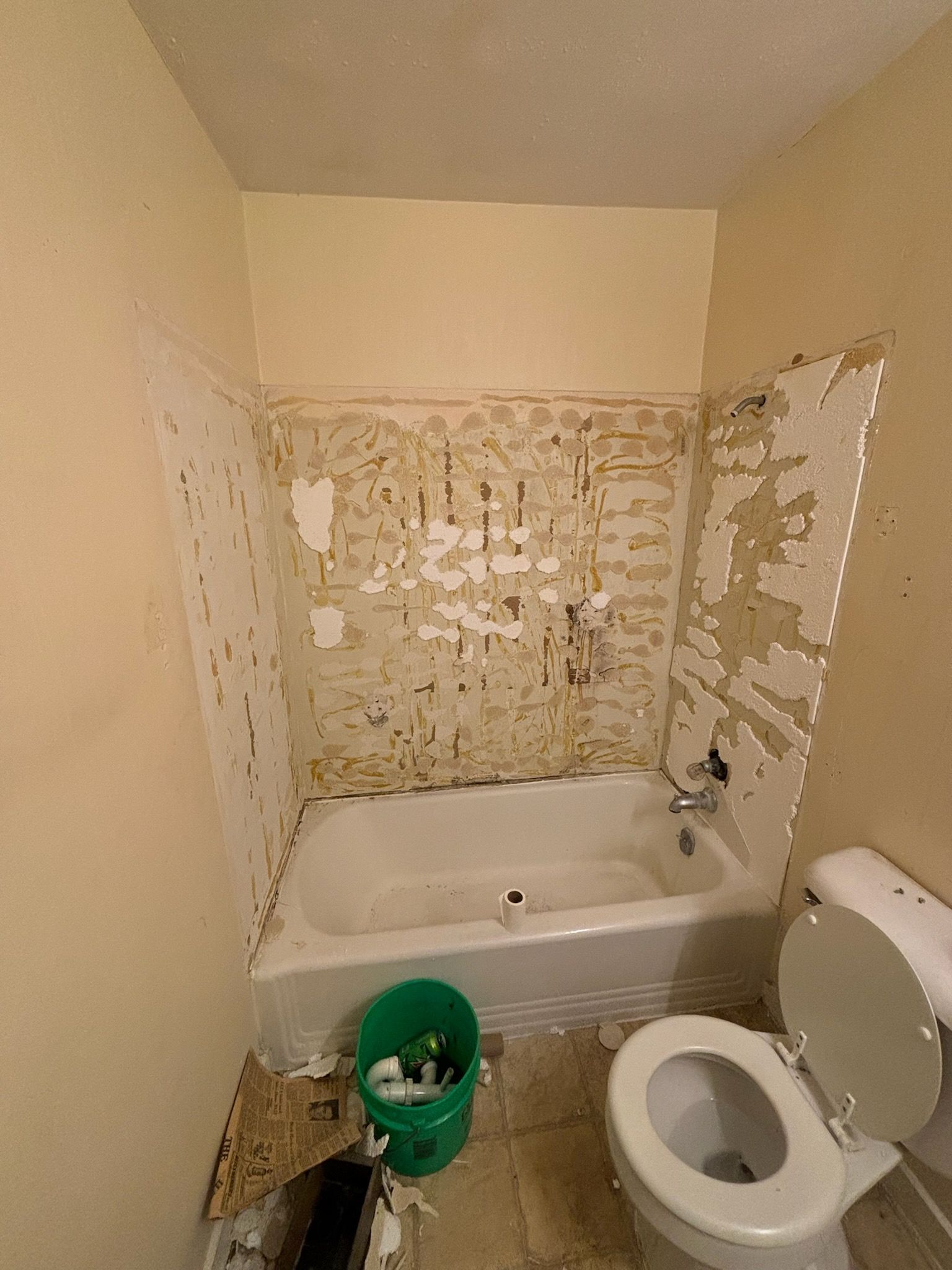 Bathroom with peeling wall around the bathtub. Toilet is on the right. Green bucket and debris on the floor.