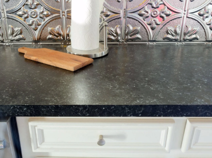 Kitchen countertop with wood cutting board, paper towel holder, and white cabinets. Silver decorative backsplash.