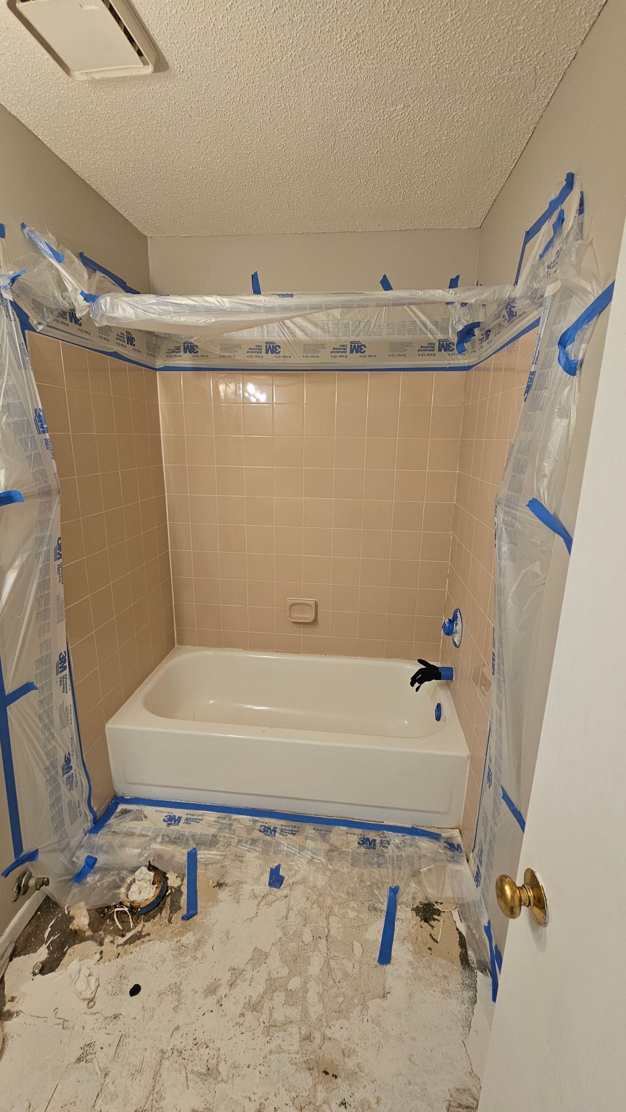 Bathroom renovation: tub and tiled walls protected with plastic and blue tape. Debris on floor.
