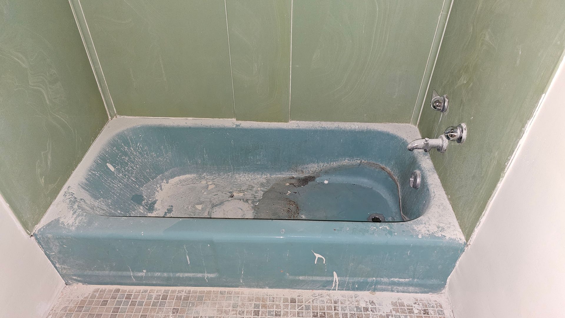 Dusty teal bathtub in a dilapidated bathroom, with green wall panels and chrome fixtures.