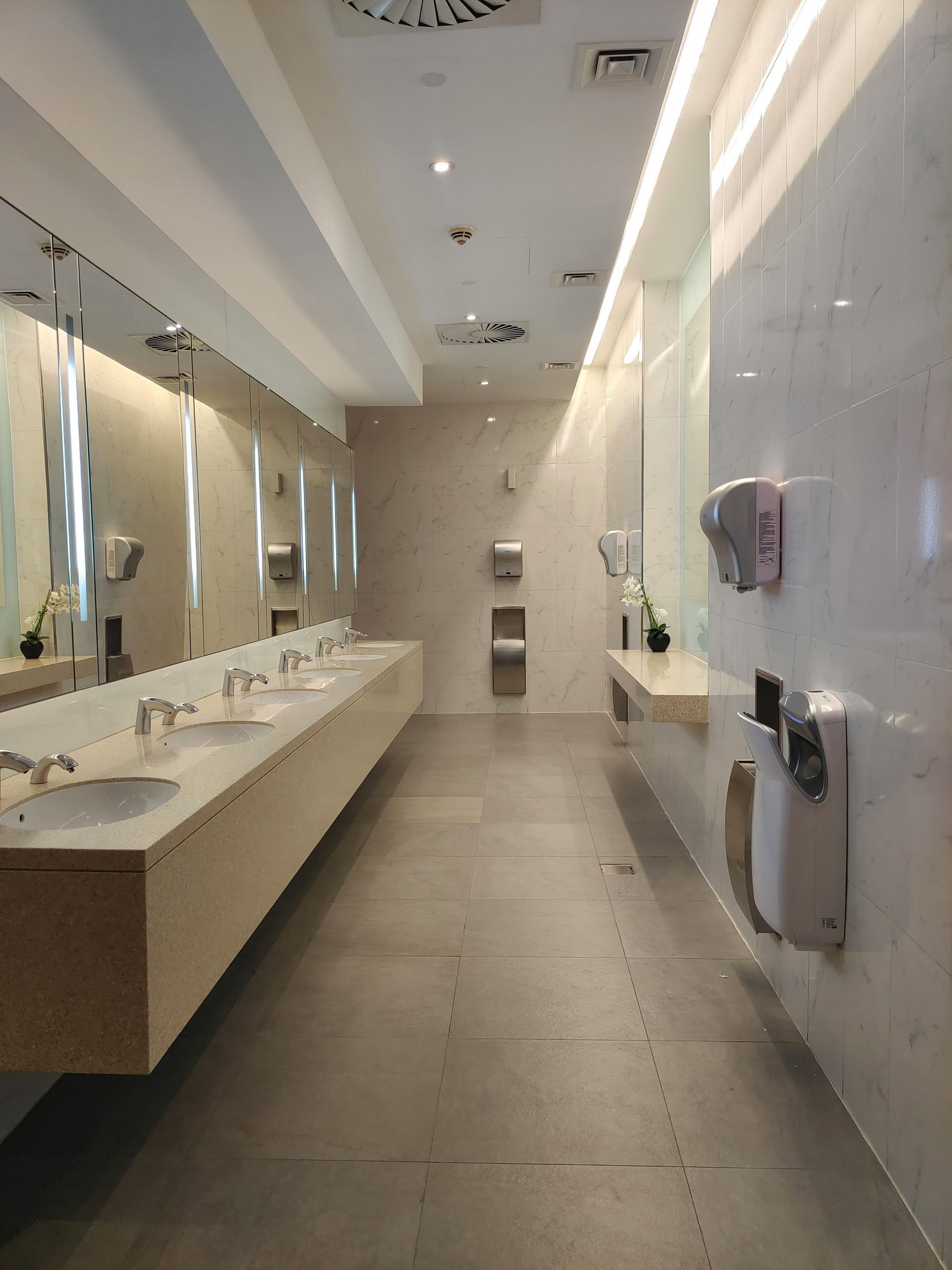Modern, well-lit public restroom with long sinks, mirrors, and hand dryers. Neutral tones and clean design.