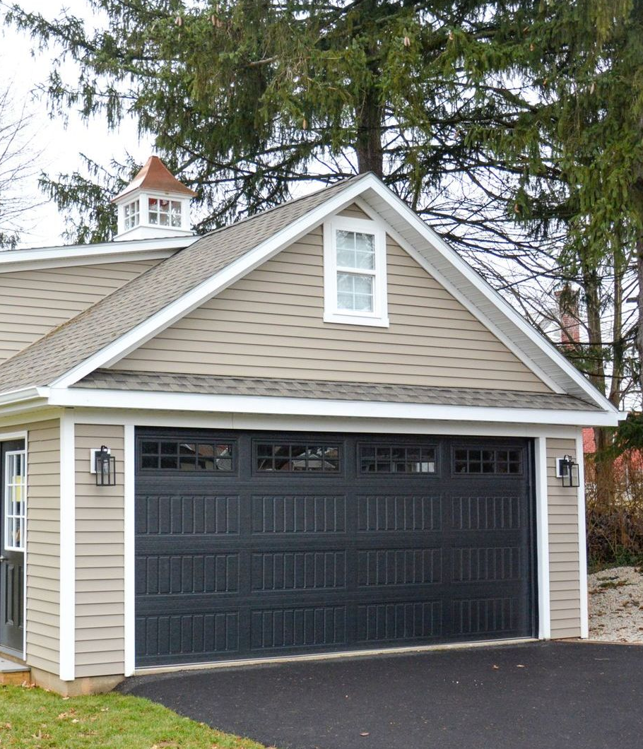 A detached garage with beige siding, a black door, a small window in the gable, and a cupola on the roof.