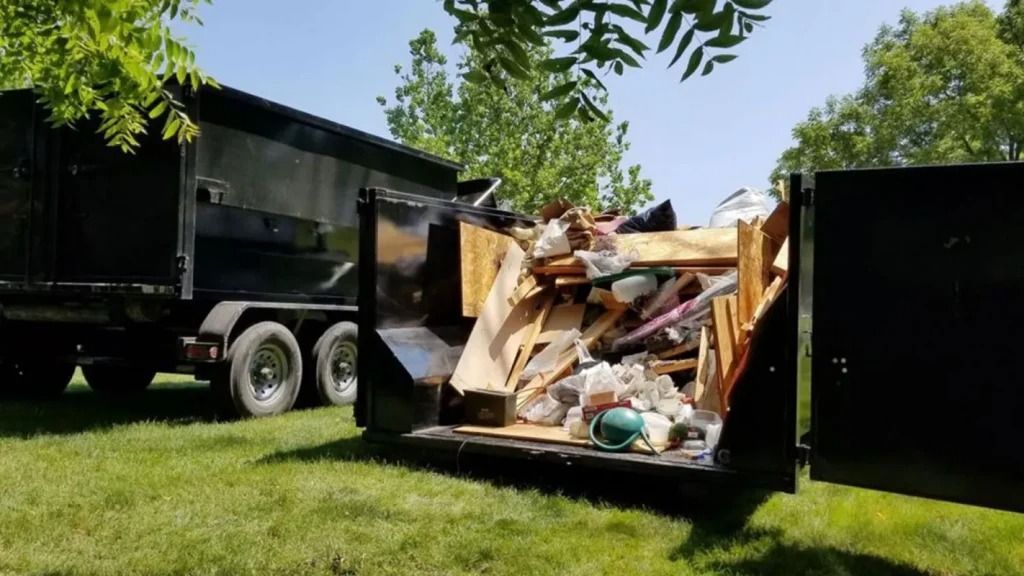 Black dumpster on wheels overflowing with construction debris, parked on grass.