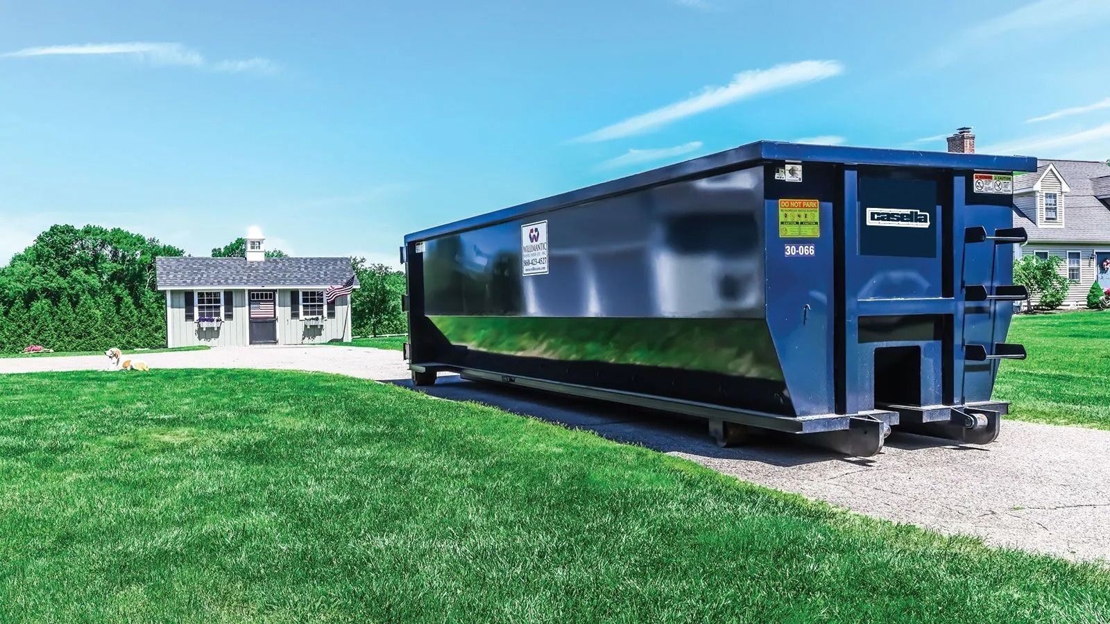 Blue dumpster on a gravel driveway, in front of a green lawn and houses under a blue sky.