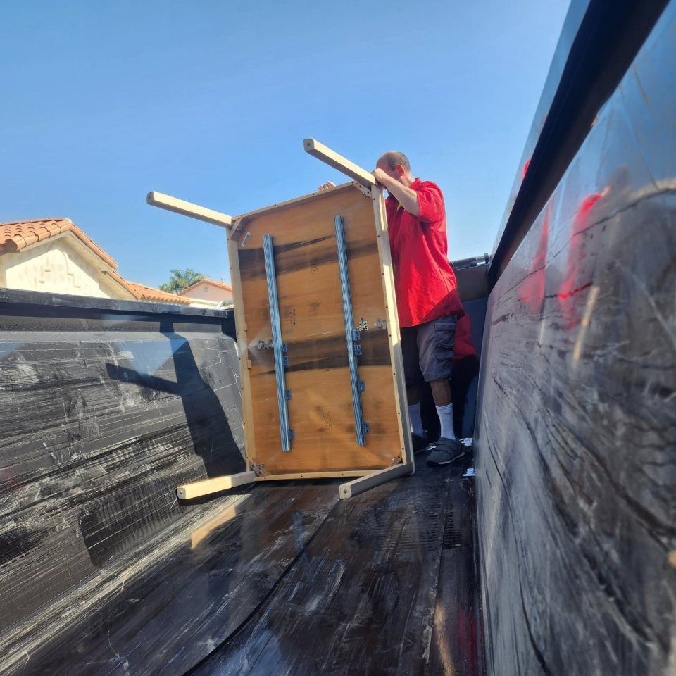 A person in a red shirt loads a wooden table into the back of a black pickup truck on a sunny day.