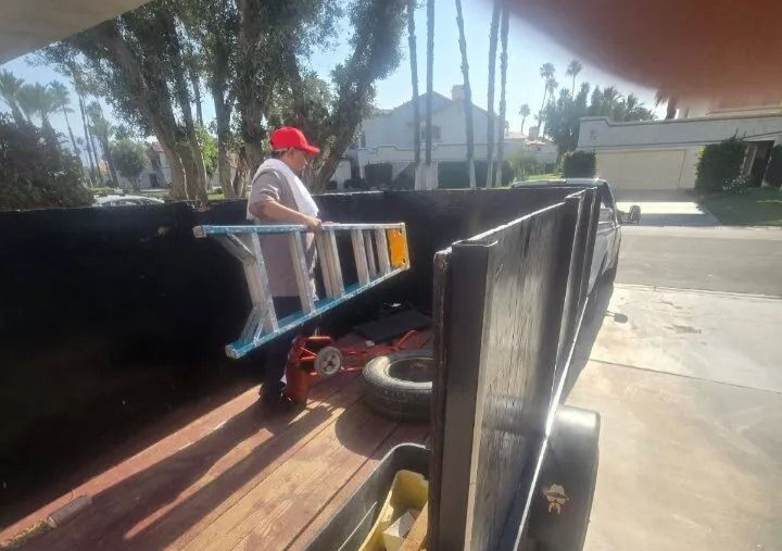 Two sanitation workers emptying a trash bin into a green truck. One directs, wearing orange safety gear.