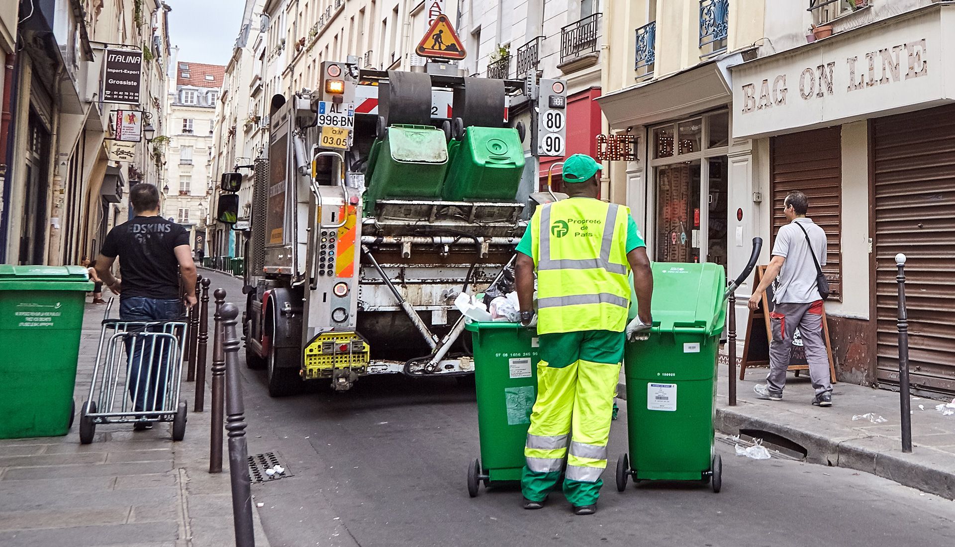 Garbage truck collecting green bins on a narrow city street. Sanitation worker in yellow, other people nearby.