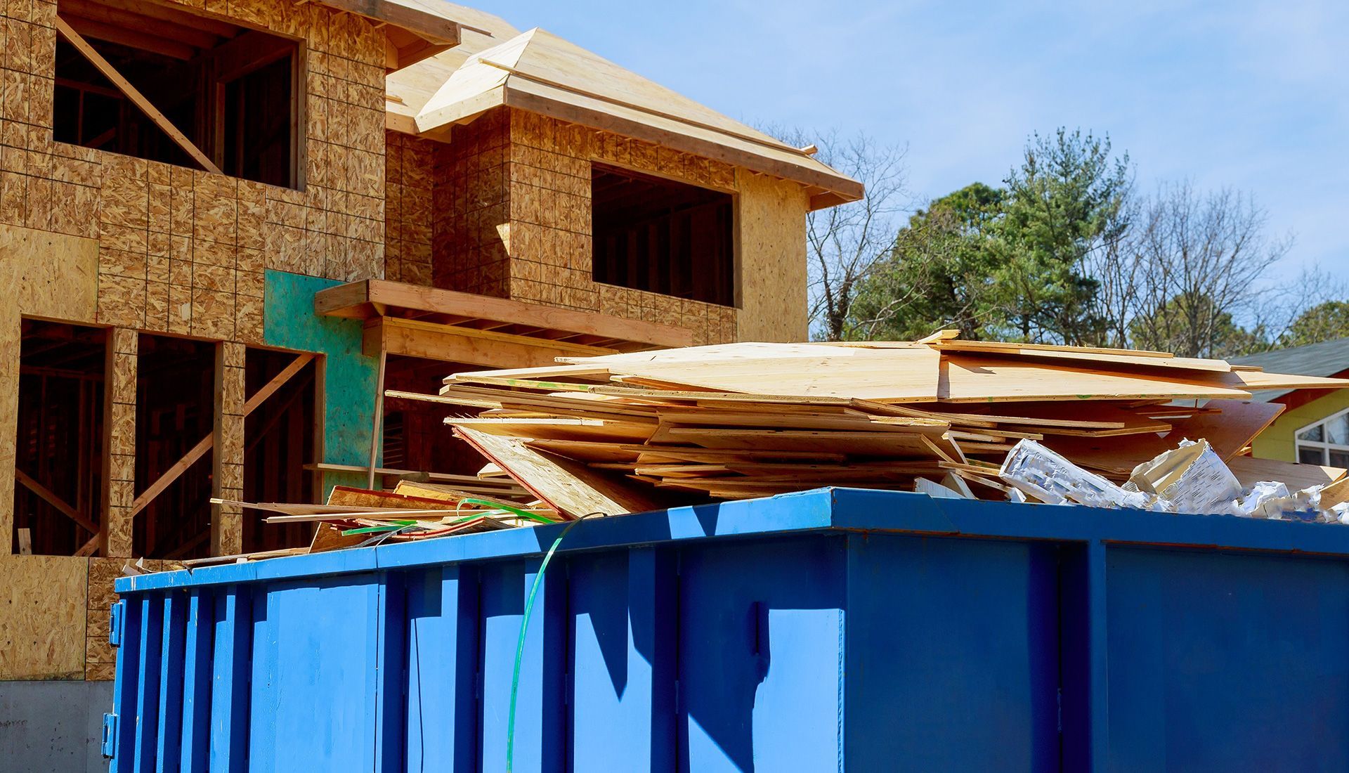 Blue dumpster filled with wood debris in front of a partially built house, sunny day.