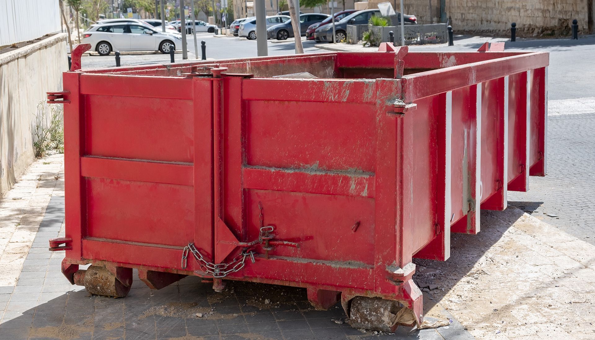 Red metal dumpster on wheels, parked on a paved street.