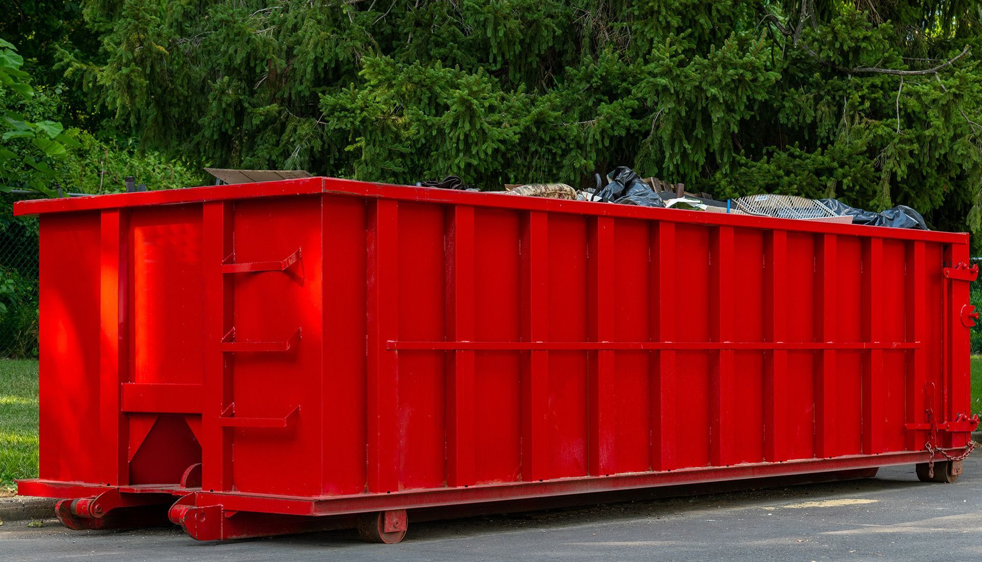 Red dumpster in a grassy area, filled with debris, with trees in the background.