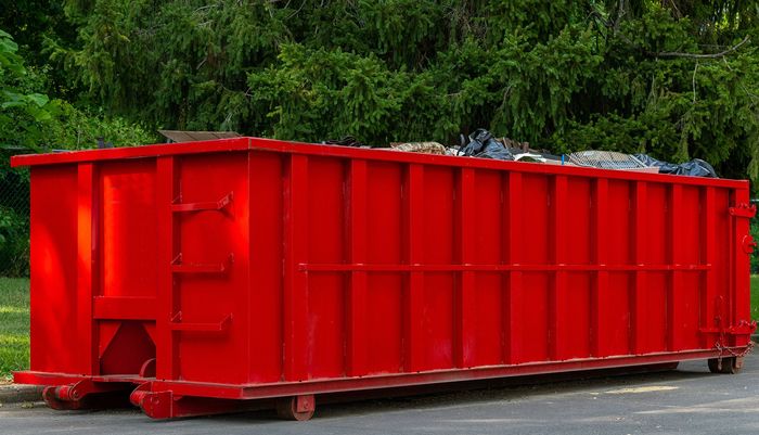 Red rectangular dumpster sits outdoors, filled with debris, with green trees in the background.