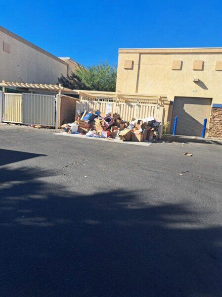 Overflowing trash bins behind a beige building. Litter and debris piled near the bins; sunny day.