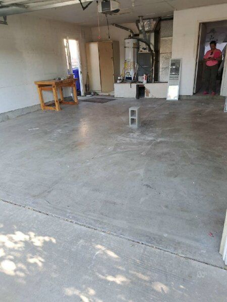A concrete-floored garage with a workbench, storage cabinet, water heater, and a person standing in the open doorway.