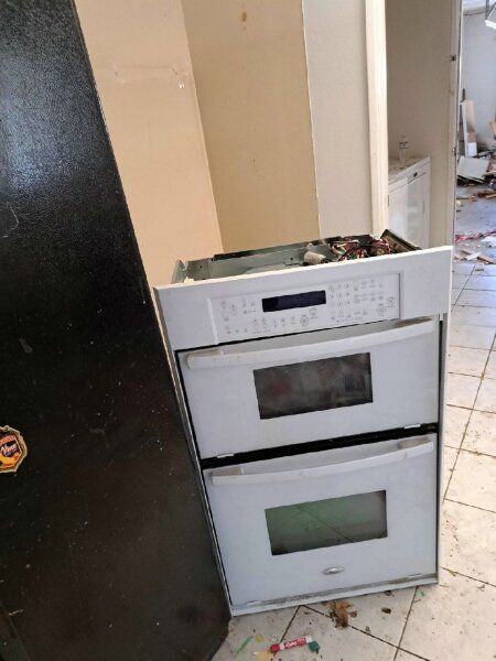 White double oven unit leaning against a black refrigerator. The oven is in a room with debris on the floor.