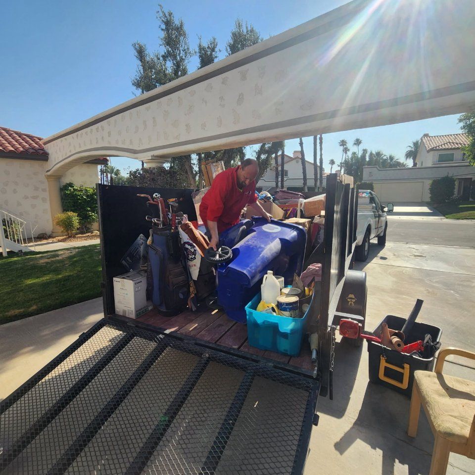 Man loads trash and junk into a trailer under a carport on a sunny day.