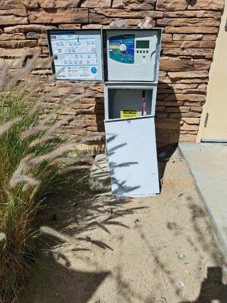 Pool equipment control box mounted on a stone wall, with open doors and weeds in the foreground.