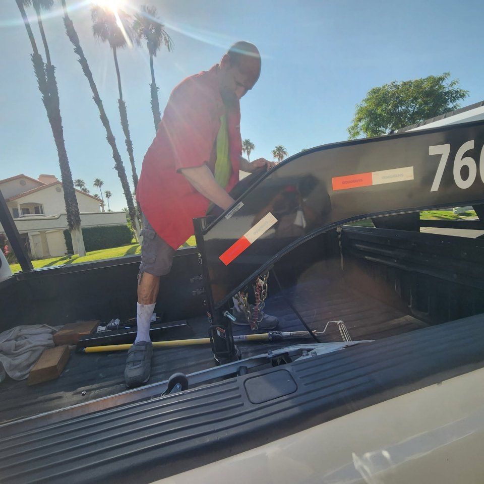 Man in red shirt working on a trailer hitch inside a truck bed on a sunny day.
