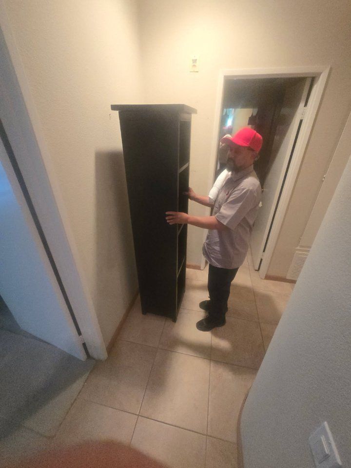 Person carrying a tall, black shelf unit in a hallway with off-white walls.